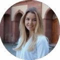 Anna Hessler lächelt freundlich in einem Portraitfoto vor einer historischen Kulisse.
